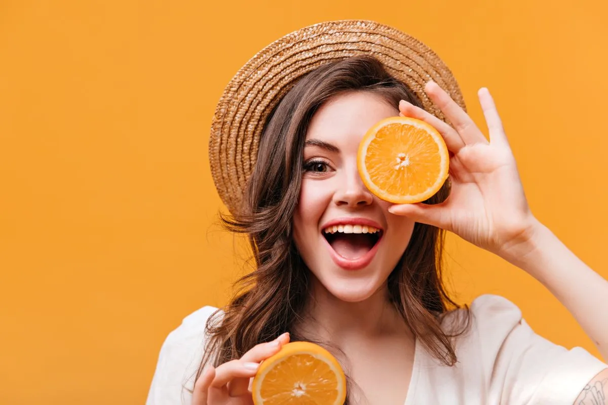 Frutas com vitamina C: quais consumir, recomendação diária e tabela 1 Frutas com vitamina C: na imagem uma mulher sorridente segura uma laranja partida em frente ao olho.